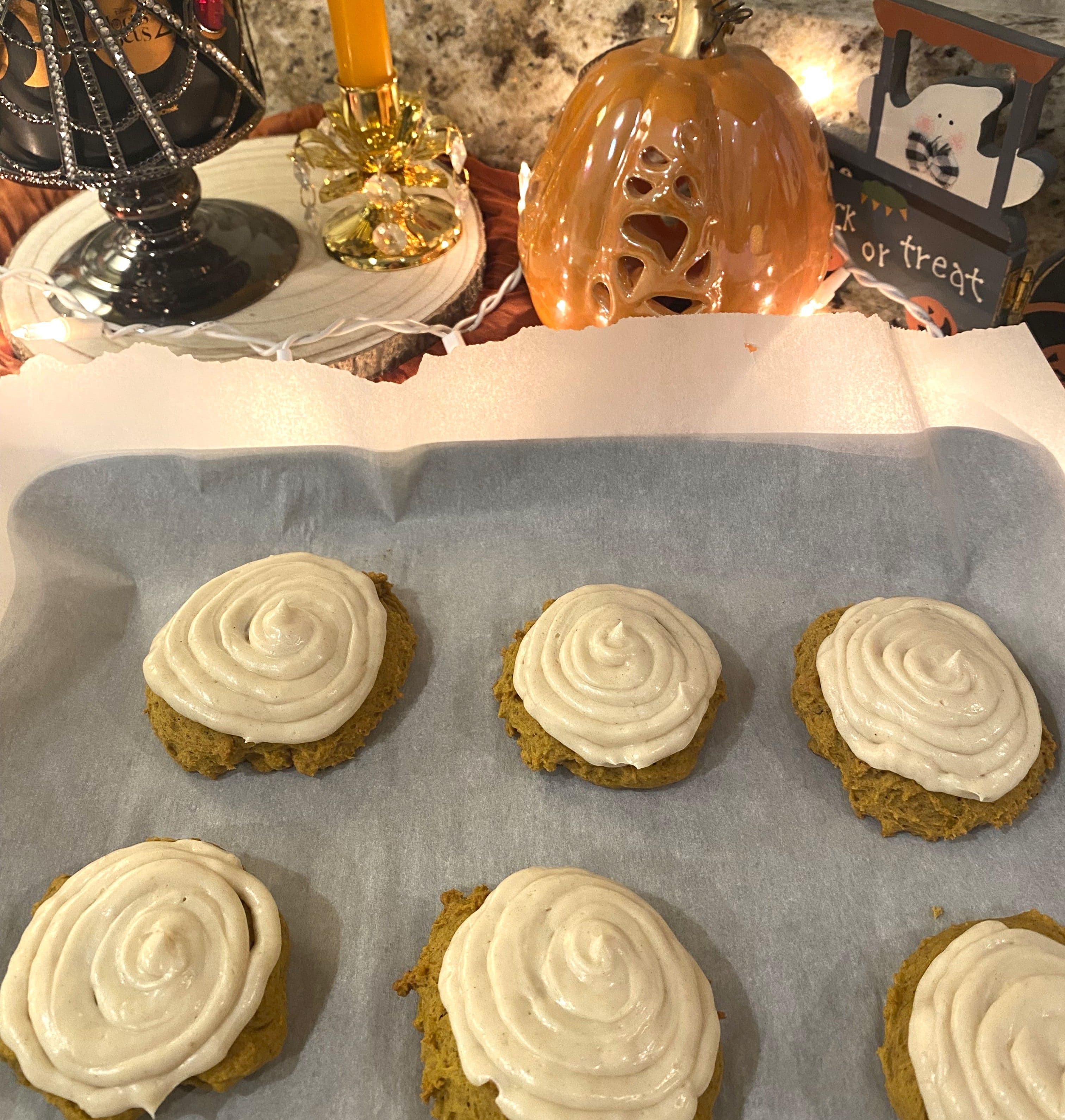 Pumpkin cookies with cream cheese frosting on baking sheet with Halloween decorations in the background.
